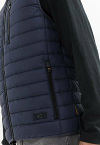 Marineblauwe gewatteerde vest met horizontaal gestikte patroon, ritszakken en een klein logo-embleem. Draagt over een donkere lange mouw shirt.