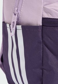 Sac à dos violet avec une surface texturée, présentant une fermeture éclair violet clair et des accents de rayures blanches sur le côté violet plus foncé.