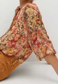 Vrouw in een blouse met puffmouwen in een rood, oranje en beige bloemenpatroon, gecombineerd met hoog getailleerde burnt orange broek.