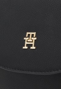 Czarna skórzana torba o fakturze z logo z złotymi elementami, które przedstawia splatające się litery "TH". Wyróżniająca się gładką i szczegółową powierzchnią.