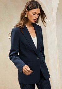 Námořnický blazer se přizpůsobeným střihem, jednořadým knoflíkovým zapínáním, klopovými reverzy a dvěma předními kapsami. Vhodné v kombinaci se světlým topem a tmavými džíny.
