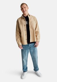 Mladý muž se usmívá, má na sobě béžovou košili s knoflíky přes černé tričko, světle modré džíny a bílé tenisky, stojí na bílém pozadí.