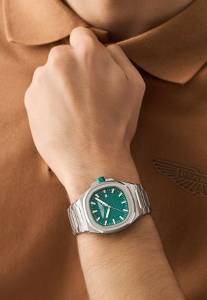 Polso con orologio in metallo argento con quadrante verde, indicatori bianchi e finestra del datario; mano che tiene il colletto di una camicia marrone con il logo Aston Martin.