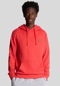 Hoodie corail lumineux avec une poche frontale, capuche à cordons et petit logo jaune sur la poitrine. Fabriqué en tissu doux et texturé.
