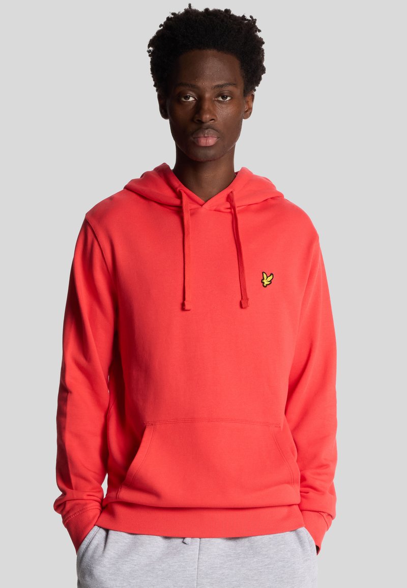 Hoodie corail lumineux avec une poche frontale, capuche à cordons et petit logo jaune sur la poitrine. Fabriqué en tissu doux et texturé.