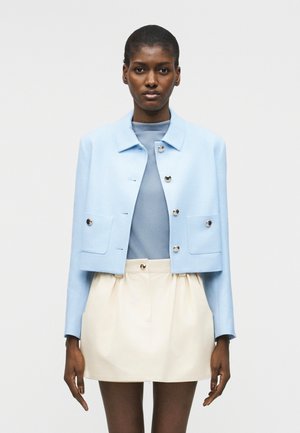 Γυναίκα που φοράει ανοιχτό μπλε κοντό σακάκι με κουμπιά, ταιριαστό τοπ και εκρού μίνι φούστα ψηλόμεση, στέκεται μπροστά σε απλό φόντο.