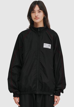 Sort zip-up jakke med røde syninger, høj krave og et logo patch på brystet. Har elastisk talje og løstsiddende ærmer.