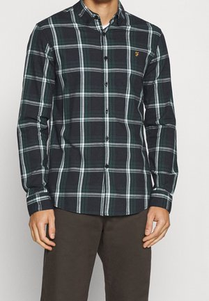 Mężczyzna w ciemnozielonej i białej kratkowanej koszuli z długim rękawem i brązowych spodniach na jednolitym tle.