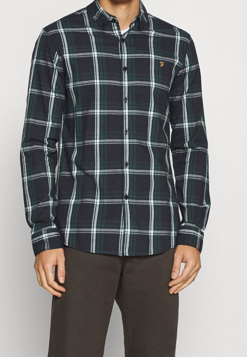 Homme portant une chemise à carreaux vert foncé et blanc à manches longues boutonnée et un pantalon marron sur un fond uni.