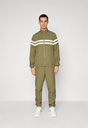 Man in olijfgroene zip-up trainingspak met witte strepen over de borst en mouwen, gecombineerd met witte sneakers, staand tegen een effen achtergrond.