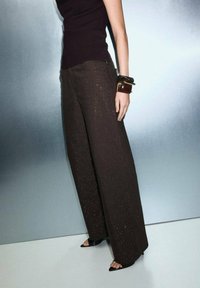 Wijde bruine broek met een gestructureerd patroon, gecombineerd met een nauwsluitend zwart topje. Een grote bruine armband en zwarte hakken zijn zichtbaar.