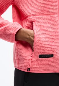 Το χέρι ενός ατόμου μέσα στην τσέπη με φερμουάρ ενός υφασμάτινου ροζ φλις μπουφάν με ορατό μαύρο λογότυπο.