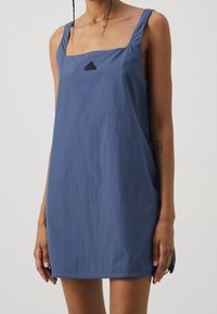 Modrá bezramenná šaty se čtvercovým výstřihem, vyrobené z lehkého materiálu. Obsahují černé logo Adidas na hrudi a postranní rozparek.
