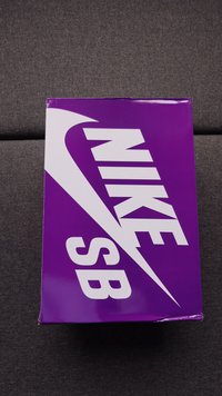 Fialová krabička na topánky Nike SB s veľkým bielym logom Nike a textom na šedej textilnej ploche.