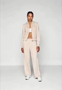 Kvinna står iförd en beige hoodie, matchande vida byxor, vit crop top och vita sneakers mot en ljus, enfärgad bakgrund.
