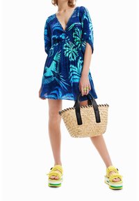 Blaues, tropisches Druckkleid mit kurzen Ärmeln und tiefem V-Ausschnitt. Accessoires bestehen aus einer geflochtenen Strohtasche und klobigen, gelben Plateausandalen.