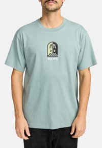 Camiseta de algodón azul claro con mangas cortas, que presenta un diseño gráfico en blanco y negro, incluyendo un patrón de tablero de ajedrez y un motivo de palmera.