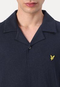 Uomo che indossa una polo blu navy con colletto aperto e un piccolo logo di aquila giallo ricamato sul petto sinistro.