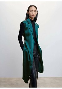 Kobieta ubrana w długą, bezrękawnikową kostium w kolorze szmaragdowej zieleni, noszącą czarny golf i czarne skórzane spodnie, stojąca na tle jasnego, jednolitego tła.