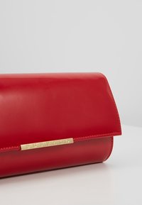 Bolso de mano de charol rojo con cierre de solapa redondeada. Presenta una placa de la marca en tono dorado en el frente y una textura lisa y brillante.