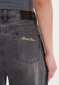 Sorte denim jeans med en baglomme, der har gylden kursiv broderi med teksten "Born Free" og en sort læderlap i livet.