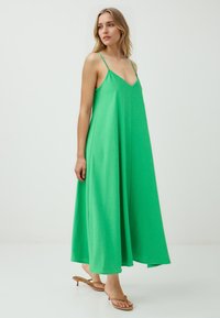 Frau in einem lebhaften grünen Maxikleid mit dünnen Trägern steht vor einem weißen Hintergrund, schaut leicht nach rechts und trägt braune Sandalen.