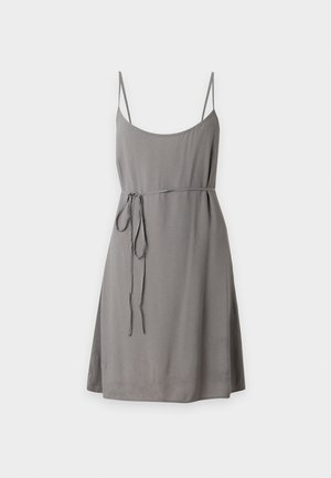 Femme aux longs cheveux foncés portant une robe grise sans manches avec une fine ceinture et des ballerines noires, debout devant un fond blanc.