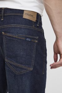 Jeans de mezclilla azul oscuro con costuras amarillas contrastantes, un parche de cuero con el nombre de la marca en la cinturilla y una etiqueta en el bolsillo trasero.