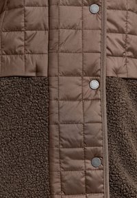 Bruin gewatteerd jack met een zachte, gestructureerde fleece paneel. Het gladde materiaal contrasteert met de harige textuur, geaccentueerd met zilveren drukknoopjes.