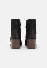 Botas de tobillo de ante negro con punta redonda, cierre de cremallera y un tacón cilíndrico marrón texturizado. Superficie suave con acentos mínimos.