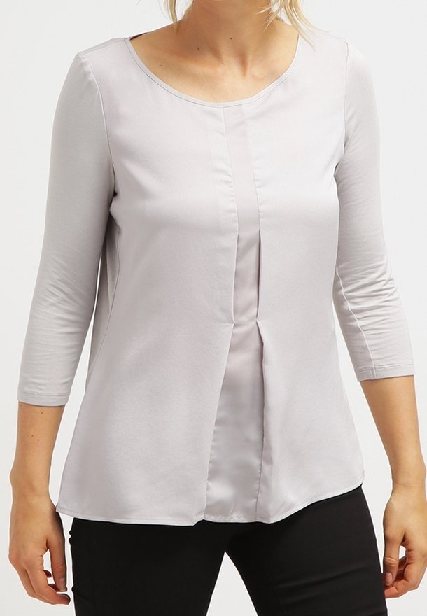 Femme portant un chemisier gris clair à manches longues avec un pli central, associé à un pantalon noir sur fond uni.