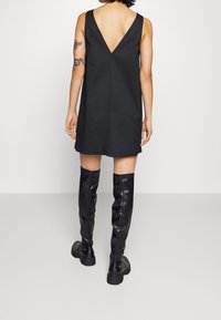 Schwarzes ärmelloses Kleid mit V-förmigem Rückenausschnitt und geradem Schnitt. Kombiniert mit glänzenden schwarzen Overknees und dicken Sohlen.