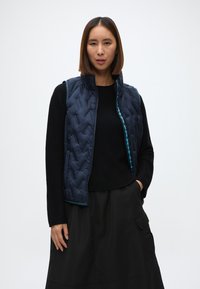 Ναυτικό γυαλιστερό γιλέκο με ψηλό γιακά, που διαθέτει κλείσιμο με φερμουάρ και αντίθετες τουρκουάζ λεπτομέρειες; φορεμένο πάνω από ένα μαύρο κορμάκι με μακριά μανίκια και φούστα.