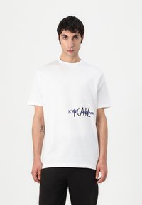 Balta medvilnės marškinėliai su apvaliu iškirpimu ir trumpomis rankovėmis, priekyje su mėlynos ir juodos spalvos „KARL“ logotipu.