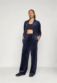 Mladá žena stojí s rukami v vreckách, oblečená v námořnicky modrom zamatovom teplákovom súprave a čiernom crop top a topánkach na svetlom pozadí.