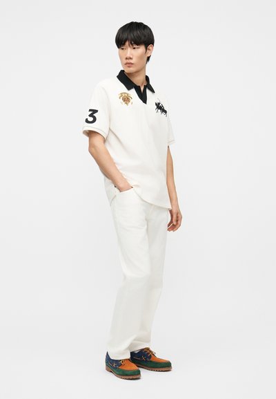 Νέος άνδρας στέκεται με τα χέρια στις τσέπες, φορώντας λευκό πόλο με μαύρο γιακά, λευκό παντελόνι και πολύχρωμα παπούτσια σε αποχρώσεις καφέ, πράσινου και μπλε.