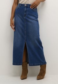 Falda maxi de denim azul oscuro con una abertura frontal, cintura alta, presillas para cinturón y bolsillos laterales. Combinada con botas de tobillo color tostado.