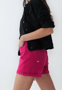 Shorts di denim fucsia con risvolti, tasche laterali e un piccolo etichetta del marchio sul retro. Abbinati a una giacca nera con bottoni.