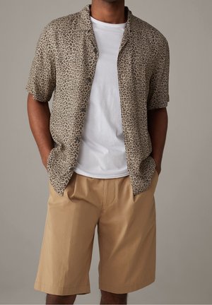 Mann trägt ein beiges Kurzarmhemd mit Leopardenmuster über einem weißen T-Shirt und hellbraune knielange Shorts, Hände in den Taschen.