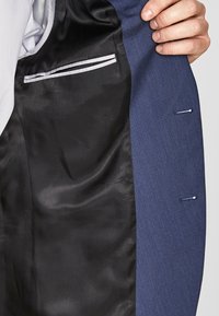 Inuti marinblazern finns en slät svart foder med en elegant ficka och ett kontrasterande texturerat blått tyg. Inkluderar stickade detaljer.