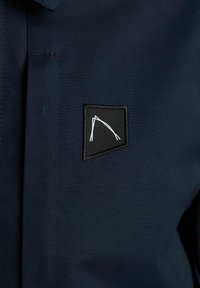 Granatowa tkanina o teksturowanej powierzchni; posiada czarną naszywkę z białym haftowanym geometrycznym wzorem.