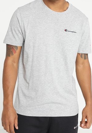 Mand iført en lysegrå Champion t-shirt og sorte shorts med Champion-logoer, der viser overkroppen og tatoverede arme.