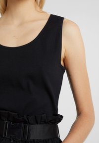 Top de tirantes negro con canalé y tirantes anchos, que presenta un escote redondeado. Detalle de cintura con volante y una hebilla negra prominente.