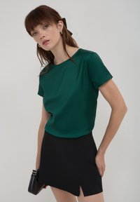 Top verde in satin a maniche corte con scollatura rotonda abbinato a una mini gonna nera con uno spacco laterale, completato con una clutch nera.