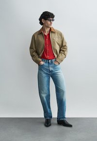 Veste marron clair avec un col contrastant, chemise rouge boutonnée, jean bleu clair et bottes noires à bout pointu, portée avec des lunettes de soleil.