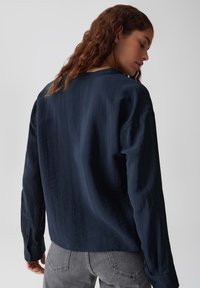 Donna con capelli castani mossi che indossa una camicia a maniche lunghe blu navy larga e jeans grigi, vista di spalle su uno sfondo grigio.