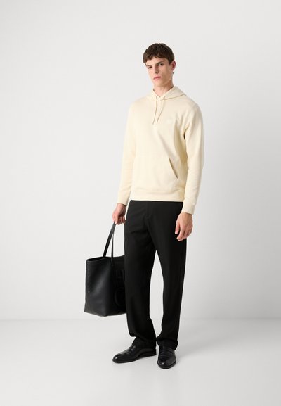 Haut à capuche crème avec poche avant et logo, associé à un pantalon noir et des chaussures noires cirées. Sac cabas noir tenu à la main droite.