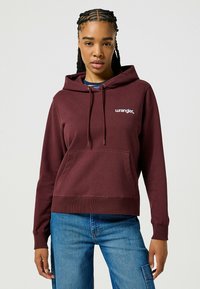 Bordeaux hoodie met een voorzak, een capuchon met trekkoord en een witte "Wrangler" logo. Heeft ribgebreide manchetten en zoom, gecombineerd met blauwe spijkerbroek.