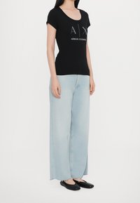 Camiseta negra de escote redondo con print de logo plateado, combinada con jeans de pierna ancha de color azul claro con dobladillo desflecado, y zapatos planos negros.