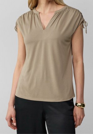 Vrouw met een kaki blouse met korte mouwen en geplooide schouders en zwarte broek, geaccentueerd met een gouden armband aan de rechterpols.